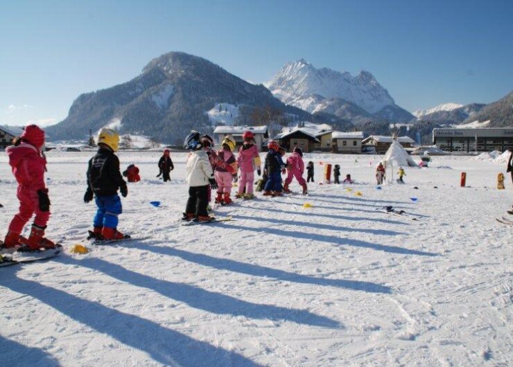 Kinder in farbenfrohen Skikostümen stehen in einer Reihe auf Schnee mit Bergen und blauem Himmel im Hintergrund.