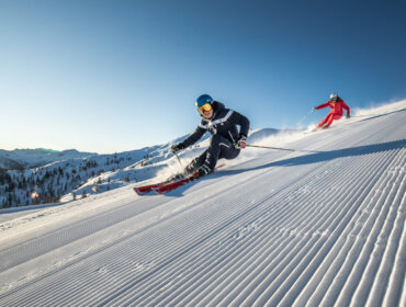 Zwei Skifahrer fahren bei strahlend blauem Himmel in den Bergen eine präparierte Schneepiste hinunter.