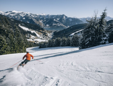 Ein Skifahrer in einer roten Jacke fährt einen verschneiten Berghang hinunter, mit malerischen Bergen und einem See im Hintergrund.