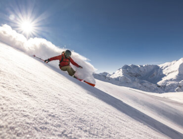 Ein Skifahrer in Rot fährt bei strahlendem Sonnenschein einen verschneiten Berghang hinunter, mit schneebedeckten Gipfeln im Hintergrund.