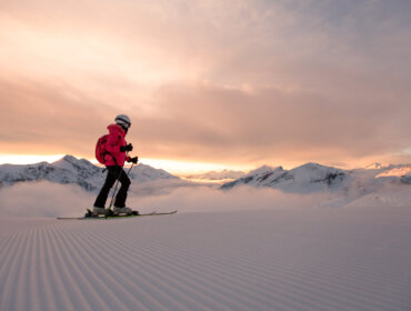 Morgenstimmung in Obertauern Eine Person fährt bei Sonnenaufgang auf präpariertem Schnee mit Bergen und Wolken im Hintergrund Ski.