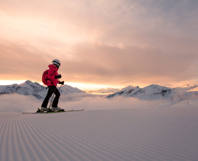 Eine Person fährt bei Sonnenaufgang auf präpariertem Schnee mit Bergen und Wolken im Hintergrund Ski.