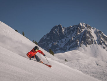 Ein Skifahrer in einer roten Jacke fährt einen verschneiten Berghang mit einem felsigen Gipfel im Hintergrund hinunter.