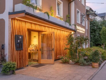 Hoteleingang mit Holzakzenten, Glastür, Pflanzen und warmer Beleuchtung am Abend.