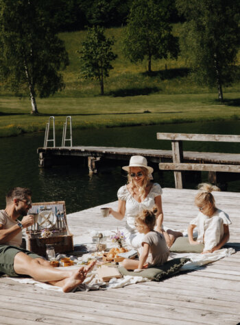 Eine vierköpfige Familie macht an einem sonnigen Tag ein Picknick auf einem hölzernen Steg an einem See, umgeben von viel Grün.