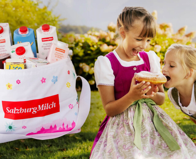 slt_0725_header_salzburg_milch_540x300 Zwei junge Mädchen in Dirndlkleidern genießen ein Gebäck im Freien neben einer mit Milchprodukten gefüllten SalzburgMilch-Tüte.