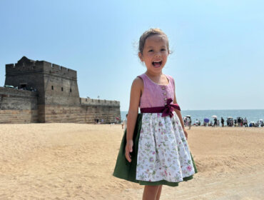 Ein lächelndes junges Mädchen in einem Kleid steht an einem Sandstrand in der Nähe einer alten Steinfestung, mit dem Meer im Hintergrund.