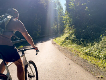 Ein Radfahrer mit Rucksack fährt auf dem sonnigen, bewaldeten und von grünen Bäumen umgebenen Alpe-Adria-Radweg bergauf.