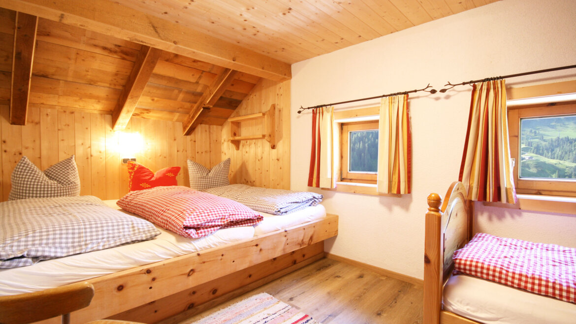 Gemütliches Almliesl-Schlafzimmer aus Holz mit zwei Betten, karierter Bettwäsche, bunten Vorhängen und einem farbenfrohen Teppich.