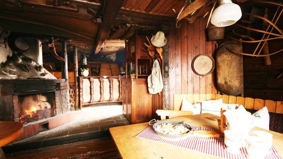 Gemütliche Almliesl-Hütte mit beleuchtetem Kamin, Holzmöbeln und alten Küchengeräten an den Wänden. (vergrößerte Ansicht)