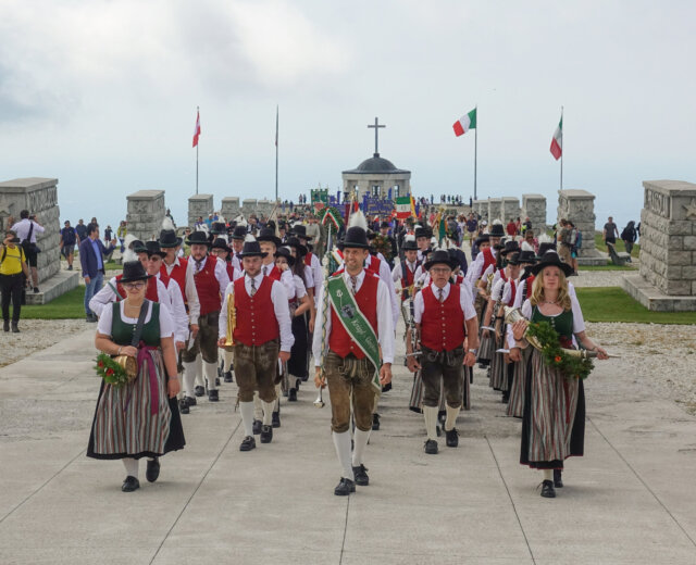 Monte-Grappa-2024_08_04-28-Kopie Menschen in traditioneller Kleidung gehen in Formation vor einem Denkmal mit Fahnen und einem Kreuz im Hintergrund.