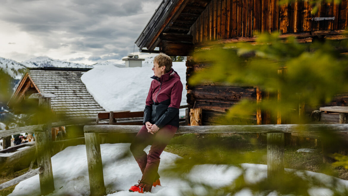 Ski-Legende Annemarie Moser-Pröll lehnt sich gegen einen Holzbalken. Im Hintergrund sieht man eine verschneite Hütte.