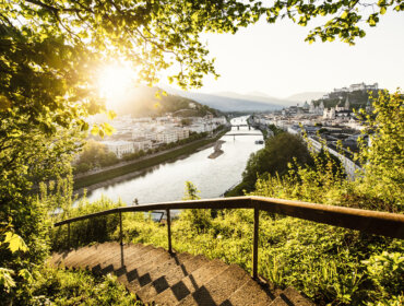Panorama Stadt Salzburg Steintreppen führen durch grünes Laub hinunter zu einer malerischen Flussstadt am Mozart-Radweg bei Sonnenuntergang, Berge dahinter.