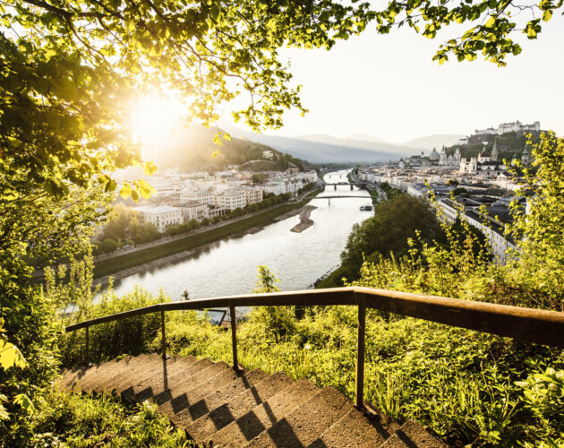 Panorama Stadt Salzburg Steintreppen führen durch grünes Laub hinunter zu einer malerischen Flussstadt am Mozart-Radweg bei Sonnenuntergang, Berge dahinter.