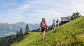 Drei Personen wandern auf einer grasbewachsenen Anhöhe mit Bergen und einer Holzhütte im Hintergrund unter blauem Himmel.