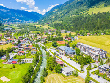 Luftaufnahme einer kleinen Stadt mit einem Fluss, grünen Feldern und Bergen unter einem blauen Himmel mit Wolken.