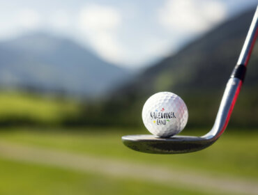 Nahaufnahme eines Golfschlägers, der einen Golfball schlägt, mit Hügeln und Himmel im Hintergrund.
