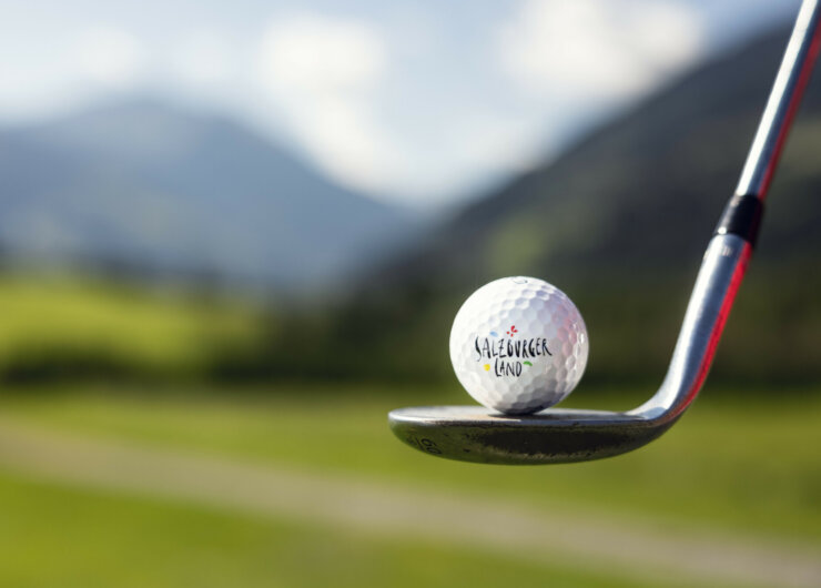 Golf Alpin – Gastein Nahaufnahme eines Golfschlägers, der einen Golfball schlägt, mit Hügeln und Himmel im Hintergrund.