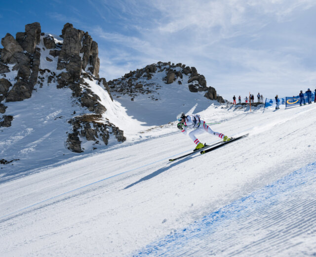 Ein Skifahrer fährt auf einem verschneiten Berghang mit felsigen Gipfeln und Zuschauern im Hintergrund bergab.