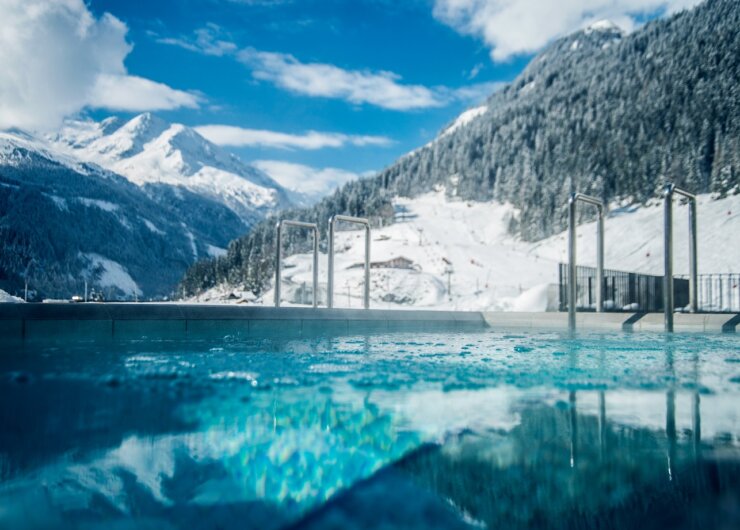 Heißer Außenpool mit sprudelndem Wasser und Blick auf verschneite Berge und einen strahlend blauen Himmel.