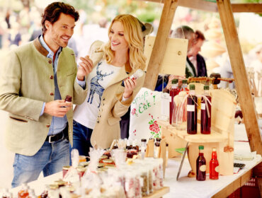 Ein lächelnder Mann und eine lächelnde Frau probieren hausgemachte Produkte an einem Marktstand mit Gläsern und Flaschen.