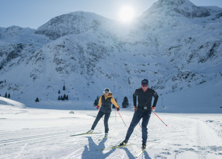 Zwei Personen beim Skilanglauf auf einer verschneiten Loipe mit Bergen im Hintergrund unter hellem Sonnenlicht.