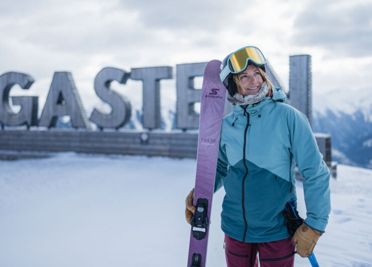 Ein lächelnder Skifahrer mit lila Skiern steht vor einem verschneiten Gasteiner Schild mit Bergen im Hintergrund.