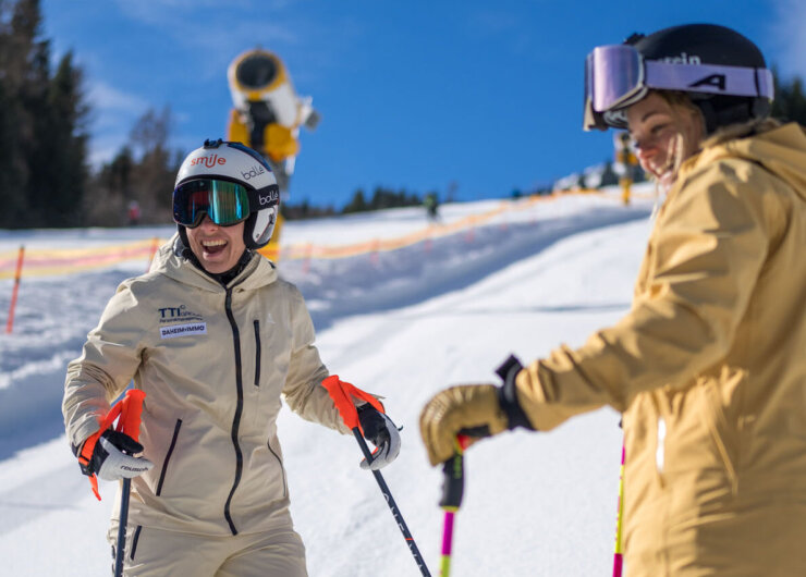 Zwei Personen in beiger Skikleidung lächeln beim Skifahren auf einer sonnigen, verschneiten Piste mit Bäumen im Hintergrund.