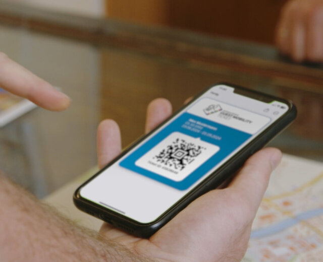 svv_24_tourismusmobilitaet_still_07-1897×1067 Eine Person hält ein Smartphone, das einen digitalen QR-Code anzeigt, über einen Tisch mit einer Karte.
