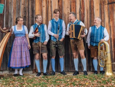 Fünf Personen in traditioneller österreichischer Kleidung, die Instrumente in der Hand halten, stehen und lächeln an einer Holzwand im Freien.