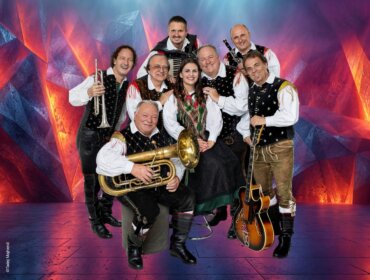 Acht Musikanten in traditioneller österreichischer Tracht posieren fröhlich mit Instrumenten vor einem farbenfrohen Hintergrund.