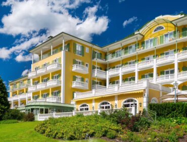 Gelbes Hotel mit weißen Balkonen und grüner Verkleidung, umgeben von Gärten unter einem strahlend blauen Himmel mit Wolken.