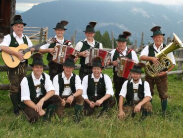 Neun Männer in traditioneller österreichischer Kleidung posieren im Freien mit Musikinstrumenten, darunter Akkordeons und eine Tuba.