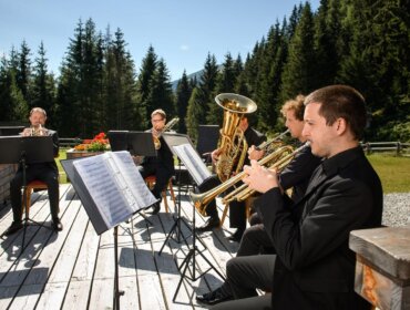 Fünf Musiker in schwarzen Anzügen spielen auf einer von Bäumen und Blumen umgebenen Holzterrasse Blasinstrumente.