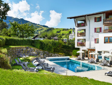 Außenpool mit Liegestühlen neben einem Hotel, eingebettet in eine grüne, malerische Berglandschaft unter blauem Himmel.