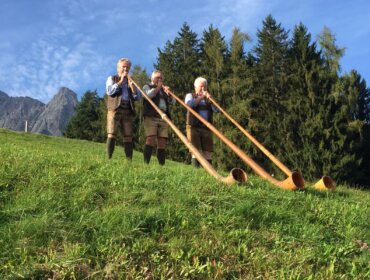 Drei Männer in traditioneller österreichischer Kleidung spielen lange Alphörner auf einem grasbewachsenen Hügel mit Bäumen und Bergen im Hintergrund.