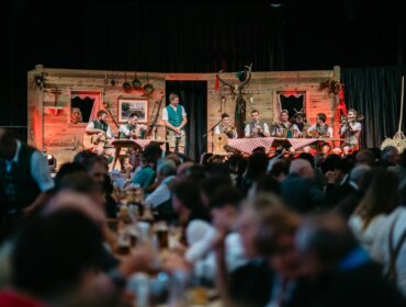 Eine Band in traditioneller Kleidung spielt auf einer rustikalen Bühne vor einer großen Menschenmenge bei einer Indoor-Veranstaltung.