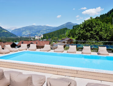 Pool auf dem Dach mit Liegestühlen und Blick auf Berge, Bäume und ein Dorf unter blauem Himmel.