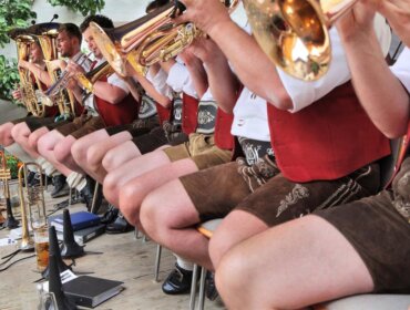 Eine Blaskapelle in österreichischen Lederhosen spielt Instrumente, in einer Reihe sitzend, mit Getränken auf dem Boden in der Nähe.