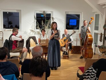 Eine Frau in österreichischer Tracht spricht zu einem Publikum, während hinter ihr eine Volksmusikgruppe mit Instrumenten sitzt.