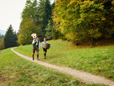 Zwei Personen in traditioneller österreichischer Kleidung gehen auf einem Waldweg und tragen eine Gitarre und ein Akkordeon.
