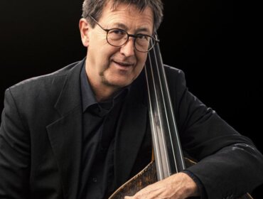 Mann mit Brille und dunklem Anzug hält einen Kontrabass vor einem schwarzen Hintergrund.