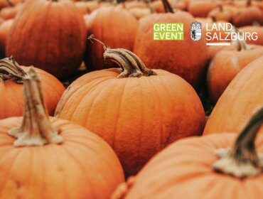 Nahaufnahme von orangefarbenen Kürbissen, mit dem Text Green Event Land Salzburg im Hintergrund.