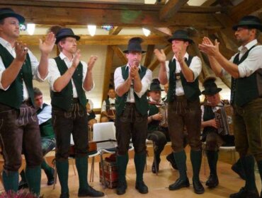 Fünf Männer in traditioneller österreichischer Kleidung klatschen, während im Hintergrund Musiker in einem hölzernen Raum spielen.