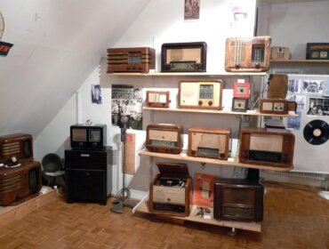 In einem Raum mit Holzfußboden und weißen Wänden stehen mehrere alte Radios auf Regalen.