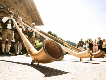 Vier Musiker spielen auf einem traditionellen Fest im Freien bei strahlendem Sonnenschein auf langen Holzalphörnern.