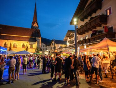 Eine Menschenmenge versammelt sich unter Zelten bei einem lebhaften Abendfest auf einem europäischen Stadtplatz.