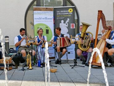 Fünf Musiker in traditioneller österreichischer Kleidung spielen im Freien mit verschiedenen Instrumenten in der Nähe von Brunnen.