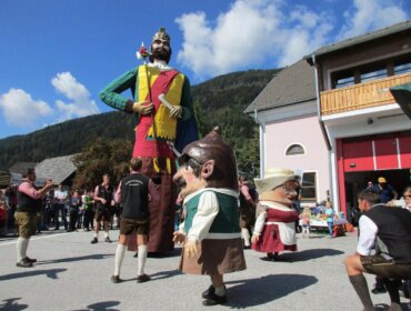 Menschen in Kostümen, darunter eine riesige Puppe und übergroße Köpfe, ziehen im Freien umher, mit den Bergen im Hintergrund.
