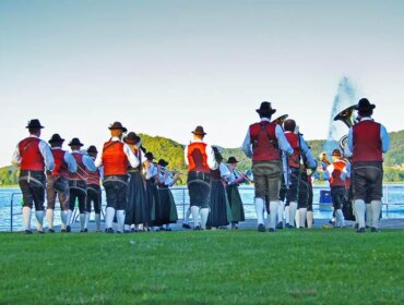 Menschen in traditioneller österreichischer Tracht treten in einer Gruppe in der Nähe eines Sees mit grünen Hügeln im Hintergrund auf.